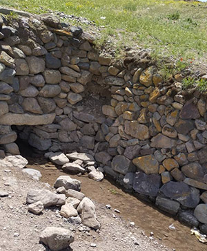 جریان باریکی از آب از دهانه‌ای سنگی در دیوار سنتی روستا بیرون می‌آید و در طبیعتی سبز جریان دارد.